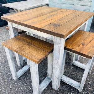 K&ouml;nnte beinhalten: Ein quadratischer Holztisch mit dunkelbraun gebeizter Platte und einem wei&szlig; gestrichenen Gestell im Used-Look, dazu zwei passende r&uuml;ckenlose B&auml;nke mit dunkelbraunen Sitzfl&auml;chen und wei&szlig; gewischten Beinen als kompaktes Essgruppen-Set.