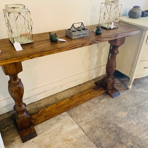 May include: A long, rectangular wooden console table with a dark brown stained finish. The table features two thick, turned pedestal legs connected by a horizontal base stretcher. The surface holds two white wire lanterns and a small egg crate.