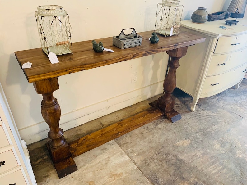 May include: A long, rectangular wooden console table with a dark brown stained finish. The table features two thick, turned pedestal legs connected by a horizontal base stretcher. The surface holds two white wire lanterns and a small egg crate.