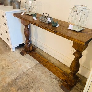 May include: A long, rectangular wooden console table with a dark brown stained finish. The table features two thick, turned pedestal legs connected by a horizontal base stretcher. The surface is flat and holds two wire lanterns and a small box.