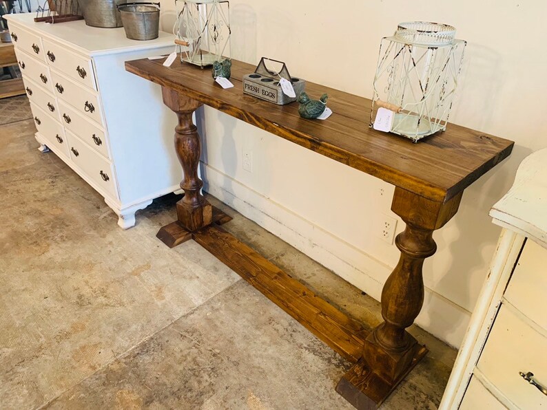 May include: A long, rectangular wooden console table with a dark brown stained finish. The table features two thick, turned pedestal legs connected by a horizontal base stretcher. The surface is flat and holds two wire lanterns and a small box.