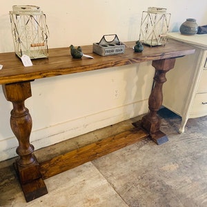 May include: A long, rectangular wooden console table with a dark brown stained finish. The table features a flat top surface supported by two thick, turned pedestal legs connected by a horizontal base stretcher near the floor.
