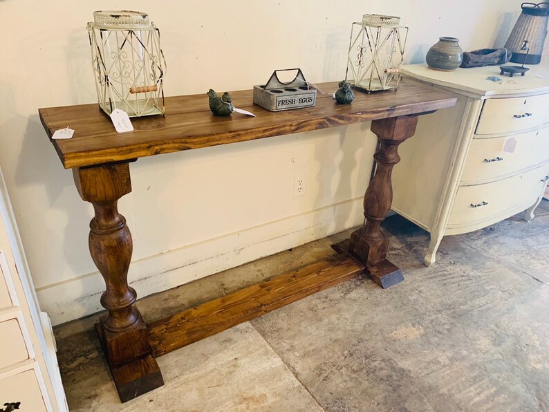 May include: A long, rectangular wooden console table with a dark brown stained finish. The table features a flat top surface supported by two thick, turned pedestal legs connected by a horizontal base stretcher near the floor.