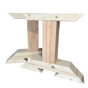 May include: Two unfinished wooden table legs. Each leg has a rectangular support base with angled edges and a central vertical support. The top horizontal support is also rectangular with angled edges. The wood grain is visible.
