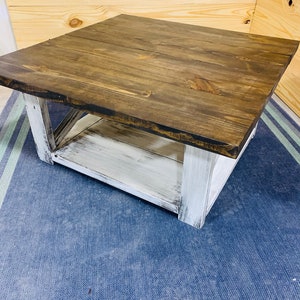 May include: A square coffee table featuring a dark brown stained wooden top with visible grain patterns. The base is constructed with four thick legs and a lower shelf, all finished in a distressed white paint that reveals wood texture.