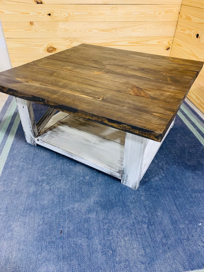 May include: A square coffee table featuring a dark brown stained wooden top with visible grain patterns. The base is constructed with four thick legs and a lower shelf, all finished in a distressed white paint that reveals wood texture.