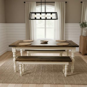 May include: A rectangular dining table with a dark brown wooden top and white turned legs, accompanied by a matching bench. The table is set with four round woven placemats and a small bowl, positioned beneath a black rectangular pendant light.
