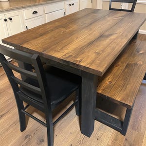 May include: A wooden dining table with a matching bench and two black chairs. The table and bench have a dark brown stained top and black legs. The chairs are black with a ladder back design. The table is approximately 72 inches long.