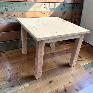 May include: A square side table constructed from light-colored natural wood with a visible grain pattern. The table features a flat, smooth top surface supported by four sturdy, straight legs positioned at each corner, showcasing a simple, functional design.
