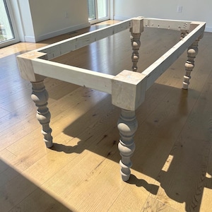 May include: A rectangular unfinished wooden table frame featuring four turned legs with a decorative bulbous design. The structure consists of light-colored raw wood beams connected at square corner blocks, showing visible pocket hole joinery.
