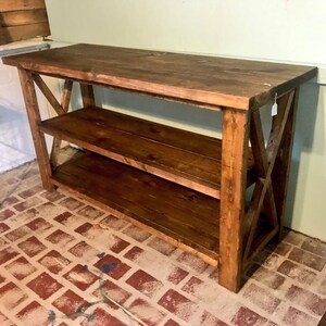 May include: A rectangular wooden console table with a dark brown stained finish. The piece features a flat top surface, one lower shelf, and side panels constructed with a prominent X-shaped support brace design on both ends of the structure.