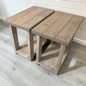 May include: Two rectangular side tables constructed from light brown wood with a visible grain pattern. Each table features a flat top surface composed of three planks, four sturdy vertical legs, and a lower shelf for additional storage space.
