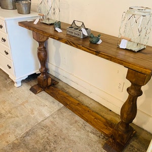 May include: A long, rectangular console table made of dark brown stained wood. The table features a flat top surface supported by two thick, turned pedestal legs connected by a horizontal base stretcher that runs along the floor between them.