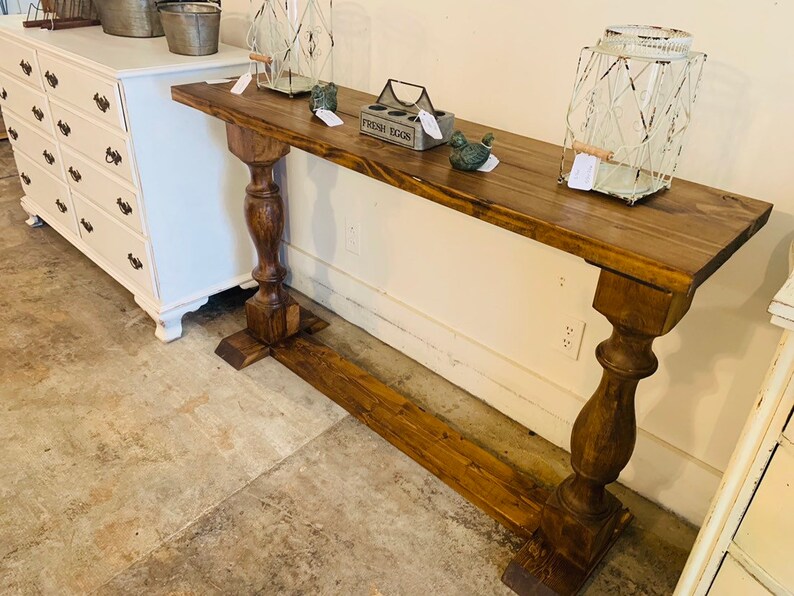 May include: A long, rectangular console table made of dark brown stained wood. The table features a flat top surface supported by two thick, turned pedestal legs connected by a horizontal base stretcher that runs along the floor between them.