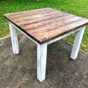 May include: A square table featuring a dark brown stained wooden top with visible wood grain and four sturdy legs finished in a distressed white paint. The table has a simple construction with a flat surface and straight, vertical legs.