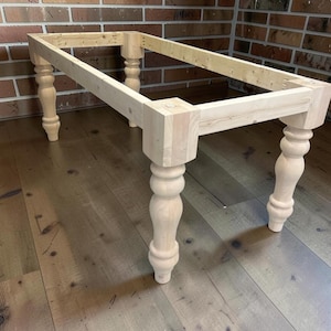 May include: Unfinished wooden table frame with four turned legs. The rectangular frame is made of light-colored wood, with visible joinery. The legs are approximately 28 inches tall, and the table is set against a brick wall.