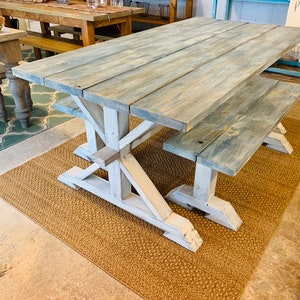 May include: A large, rectangular farmhouse-style dining table with a matching bench. The tabletop and bench are made of light blue-gray stained wood. The base is white with an X-shaped support. The table is 72 inches long.