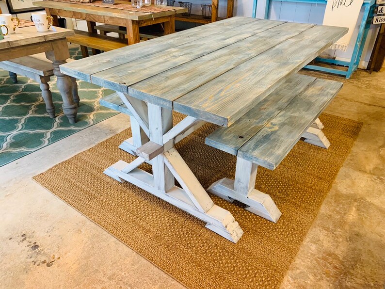 May include: A large, rectangular farmhouse-style dining table with a matching bench. The tabletop and bench are made of light blue-gray stained wood. The base is white with an X-shaped support. The table is 72 inches long.