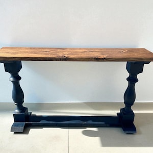May include: A console table featuring a rectangular wooden top with a natural brown wood grain finish. The table is supported by two black painted pedestal legs with a turned design, connected by a horizontal black base stretcher at the bottom.