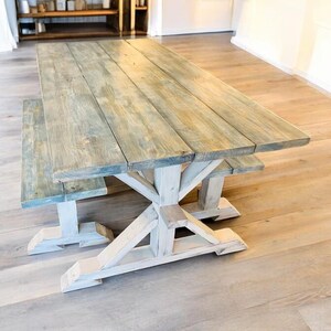 May include: A rustic wooden dining table and bench set. The tabletop has a weathered blue-gray finish, constructed from planks. The base and legs are painted white, featuring an X-shaped design. The set is designed for indoor use.
