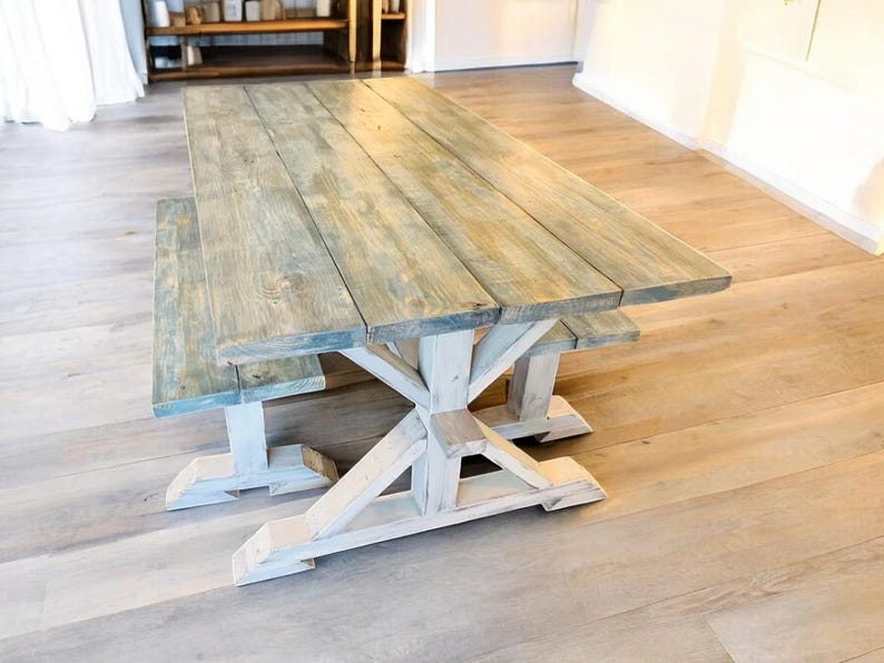 May include: A rustic wooden dining table and bench set. The tabletop has a weathered blue-gray finish, constructed from planks. The base and legs are painted white, featuring an X-shaped design. The set is designed for indoor use.