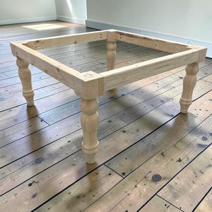 May include: Unfinished wooden table frame with four turned legs. The rectangular frame is made of light-colored wood, with visible wood grain. The legs are a classic turned design, adding a touch of elegance. The table frame is ready for finishing and assembly.