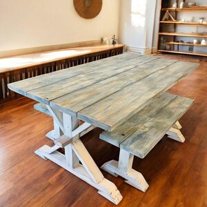 May include: A rustic wooden dining table and bench set. The tabletop and bench have a weathered, gray-blue finish, while the trestle-style base is painted white. The set is made of wooden planks and is suitable for indoor use.