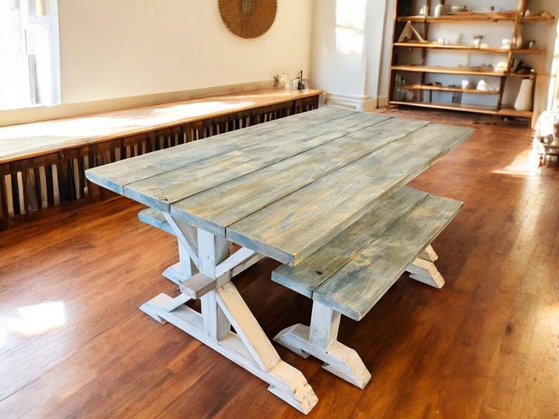 May include: A rustic wooden dining table and bench set. The tabletop and bench have a weathered, gray-blue finish, while the trestle-style base is painted white. The set is made of wooden planks and is suitable for indoor use.