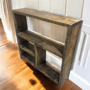 May include: A wall-mounted wooden shelf unit featuring a dark brown stained finish with visible wood grain. The structure includes a long top surface, a full-width middle shelf, and a lower section divided into two separate compartments by a vertical divider.