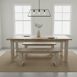 May include: A rectangular light-toned wooden dining table with two matching benches. The table is set with two white plates and a central beige vase containing green foliage. A black metal three-light pendant fixture hangs above the table.