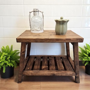 May include: A dark brown wooden table with a shelf. The table has a rustic look and is made of reclaimed wood. There are two plants on either side of the table, and a white lantern and a green ceramic jar on top of the table.