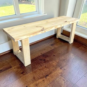May include: A light-colored wooden bench featuring a rectangular seat with visible wood grain and knots. The bench is supported by two sturdy, U-shaped leg structures made of matching light-toned wood, providing a stable base for the seat.