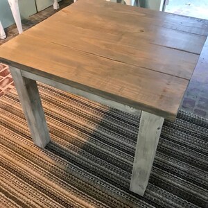 May include: A square wooden table with a light brown stain and white painted legs. The table is sitting on a striped rug.