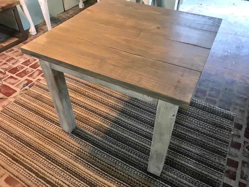 May include: A square wooden table with a light brown stain and white painted legs. The table is sitting on a striped rug.