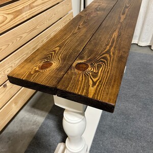 May include: A console table featuring a long, rectangular top made of two dark brown stained wooden planks with visible wood grain and knots. The tabletop is supported by a white painted pedestal base with a turned, spindle-style column.
