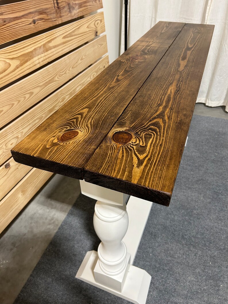 May include: A console table featuring a long, rectangular top made of two dark brown stained wooden planks with visible wood grain and knots. The tabletop is supported by a white painted pedestal base with a turned, spindle-style column.