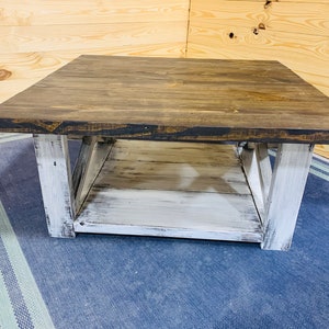 May include: A square coffee table featuring a dark brown stained wooden top and a lower shelf. The base and legs are finished in a distressed white paint, showcasing a visible wood grain texture and X-shaped side supports for structural stability.