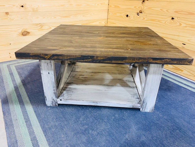 May include: A square coffee table featuring a dark brown stained wooden top and a lower shelf. The base and legs are finished in a distressed white paint, showcasing a visible wood grain texture and X-shaped side supports for structural stability.