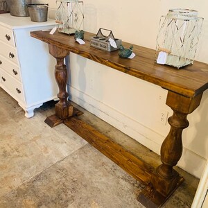 May include: A long, narrow console table with a dark brown wood finish. The table features a rectangular top supported by two thick, turned pedestal legs connected by a horizontal base stretcher. The surface holds two wire lanterns and a metal egg crate.