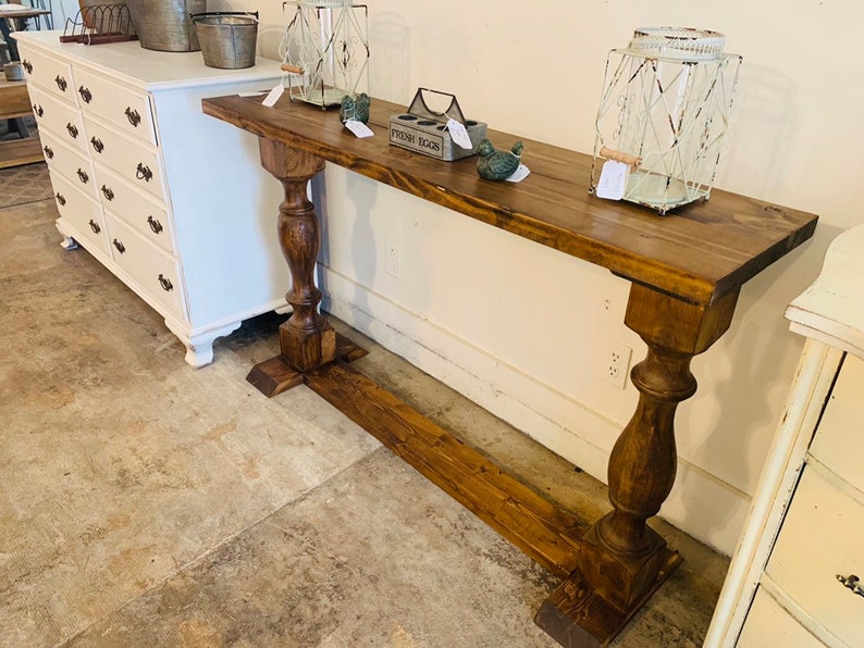 May include: A long, narrow console table with a dark brown wood finish. The table features a rectangular top supported by two thick, turned pedestal legs connected by a horizontal base stretcher. The surface holds two wire lanterns and a metal egg crate.