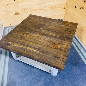 May include: A square coffee table featuring a dark brown stained wooden top with visible wood grain and knots. The base of the table is constructed from white painted wood with a distressed finish, showing the underlying texture of the material.
