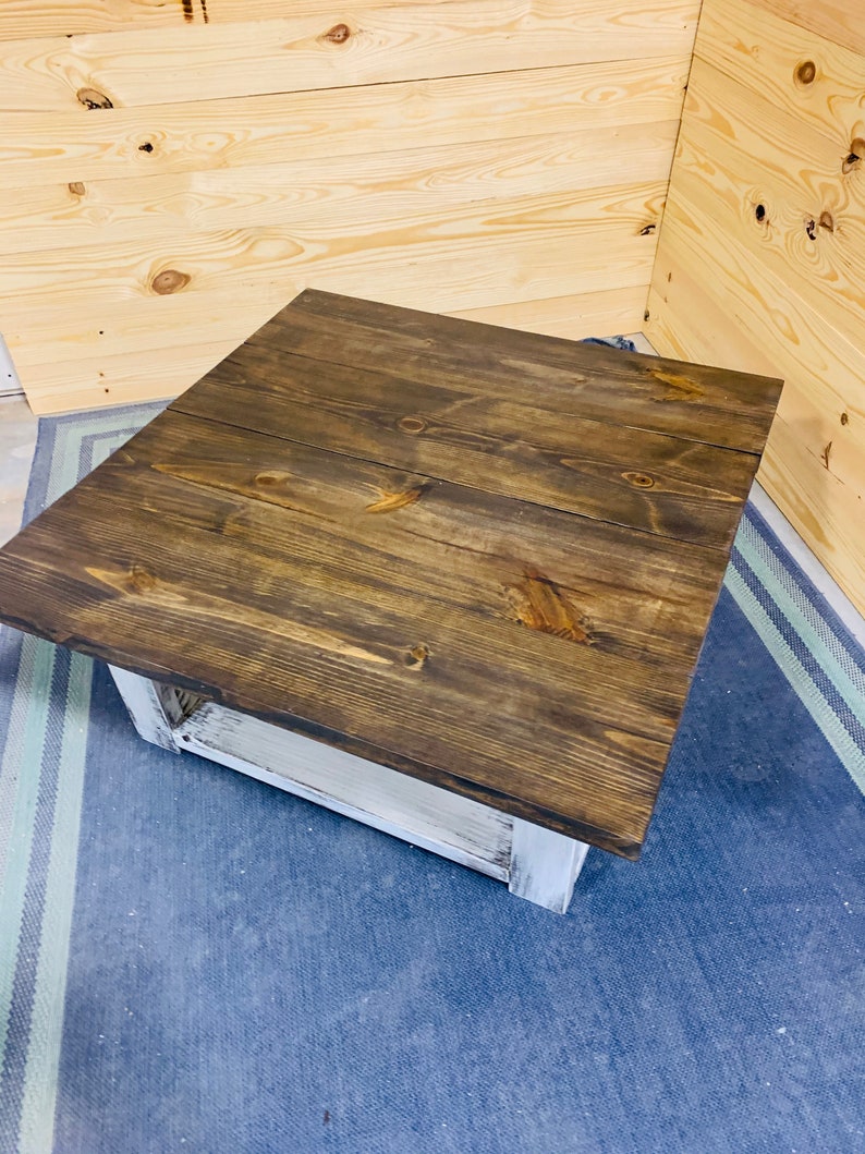 May include: A square coffee table featuring a dark brown stained wooden top with visible wood grain and knots. The base of the table is constructed from white painted wood with a distressed finish, showing the underlying texture of the material.