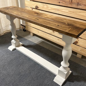 May include: A long rectangular console table featuring a dark brown stained wooden top with visible wood grain patterns. The table is supported by two white painted baluster-style legs connected by a horizontal white base stretcher piece.
