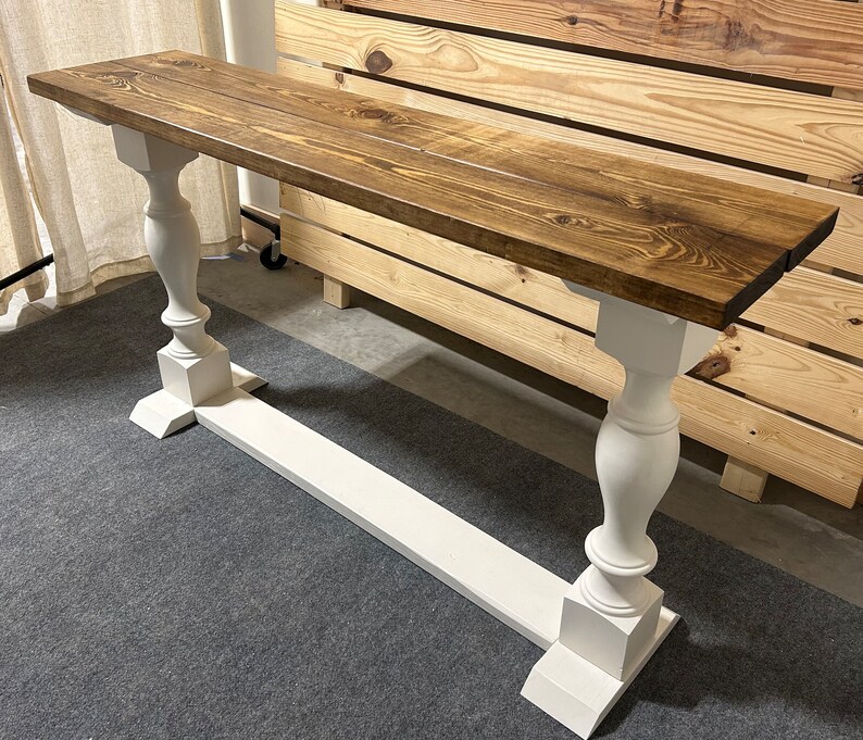 May include: A long rectangular console table featuring a dark brown stained wooden top with visible wood grain patterns. The table is supported by two white painted baluster-style legs connected by a horizontal white base stretcher piece.