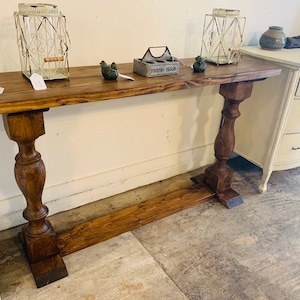 May include: A long, rectangular wooden console table with a dark brown stained finish. The table features a flat top surface supported by two thick, turned pedestal legs connected by a horizontal base stretcher near the floor.