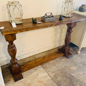 May include: A long, rectangular wooden console table with a dark brown stained finish. The table features a flat top surface supported by two thick, turned pedestal legs connected by a horizontal base stretcher near the floor.
