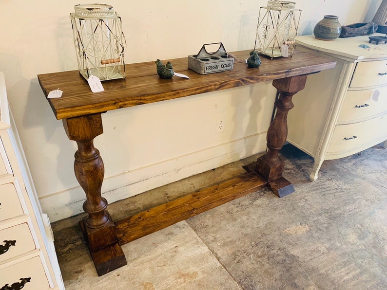May include: A long, rectangular wooden console table with a dark brown stained finish. The table features a flat top surface supported by two thick, turned pedestal legs connected by a horizontal base stretcher near the floor.