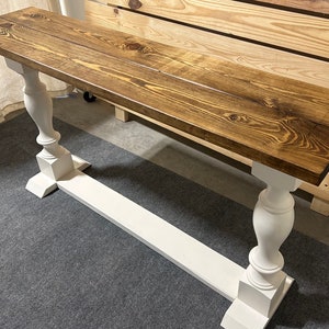 May include: A rectangular console table featuring a dark brown stained wooden top with visible wood grain patterns. The table is supported by two white painted pedestal legs connected by a horizontal base stretcher, creating a stable structure.