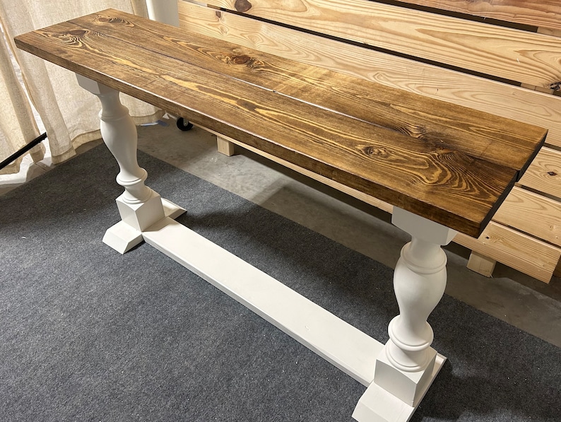 May include: A rectangular console table featuring a dark brown stained wooden top with visible wood grain patterns. The table is supported by two white painted pedestal legs connected by a horizontal base stretcher, creating a stable structure.