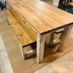 May include: A rustic wooden dining table and bench set. The table and bench feature a natural wood finish with visible grain and knots. The legs are painted white, and the set sits on a woven jute rug.